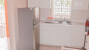 Cocina compacta con armarios blancos, refrigerador de acero inoxidable y una ventana con celosías decorativas.