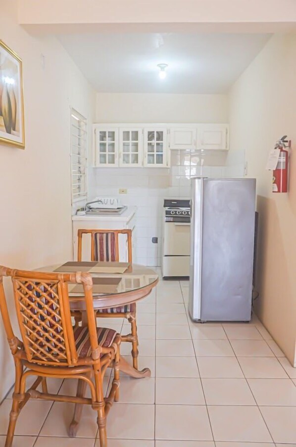 A compact kitchen with white cabinets, a stainless steel refrigerator, and a dining area with a wooden table and chairs. - Urban Apartments (Runaway Bay)