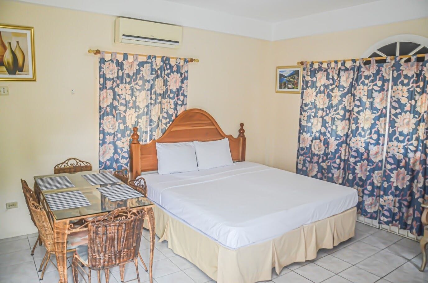 A bedroom with a wooden bed, a checkered table, wicker chairs, floral curtains, and a tiled floor.