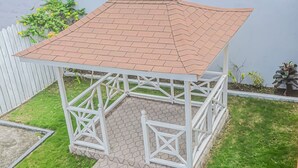 Un pabellón con techo de tejas y estructura metálica blanca, rodeado de un pequeño jardín y una valla blanca.