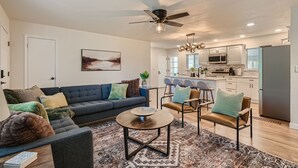 A modern living room with a dark blue sofa, a round coffee table, and a patterned rug. There is a ceiling fan, a kitchen with white cabinets, and a refrigerator.