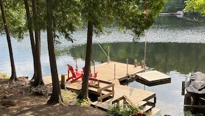 A wooden dock extending into a calm lake, with red Adirondack chairs and a covered grill area.