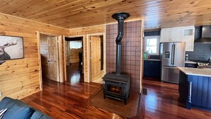 A wooden interior with a central wood stove, a kitchen with modern appliances, and a hallway leading to another room.