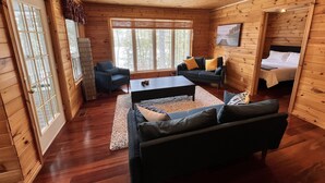 A cozy cabin with wooden walls and floors, a large window, a sofa, a coffee table, and a bed.