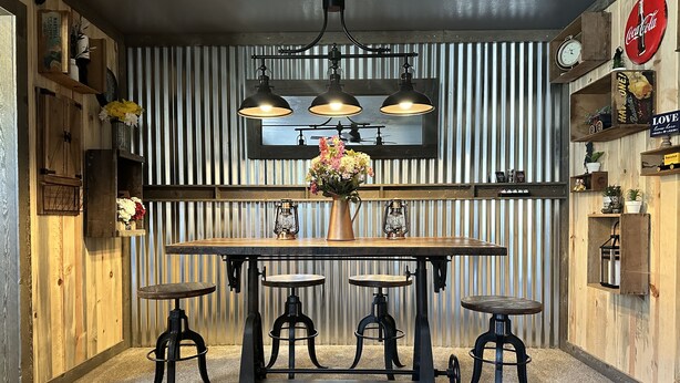 Une cuisine rustique avec une table en bois, quatre tabourets, un luminaire suspendu et une fenêtre équipée de stores verticaux.