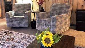 Un salon avec deux fauteuils à motifs, une table basse ornée d’un vase de tournesols, un mur en bois avec une horloge et une lampe.