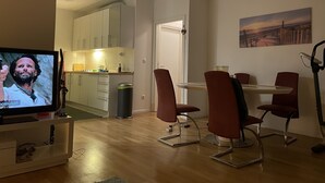 Una sala de estar moderna con televisor, comedor con sillas rojas, cocina con alacenas blancas y un cuadro en la pared.