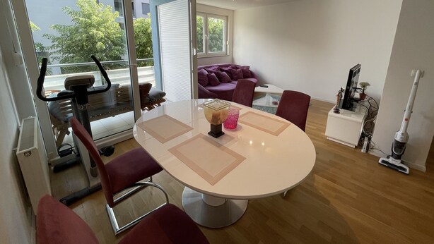 Una sala de estar moderna con un ventanal que da a un balcón, una mesa de comedor redonda con sillas, un sofá morado y una cinta de correr.