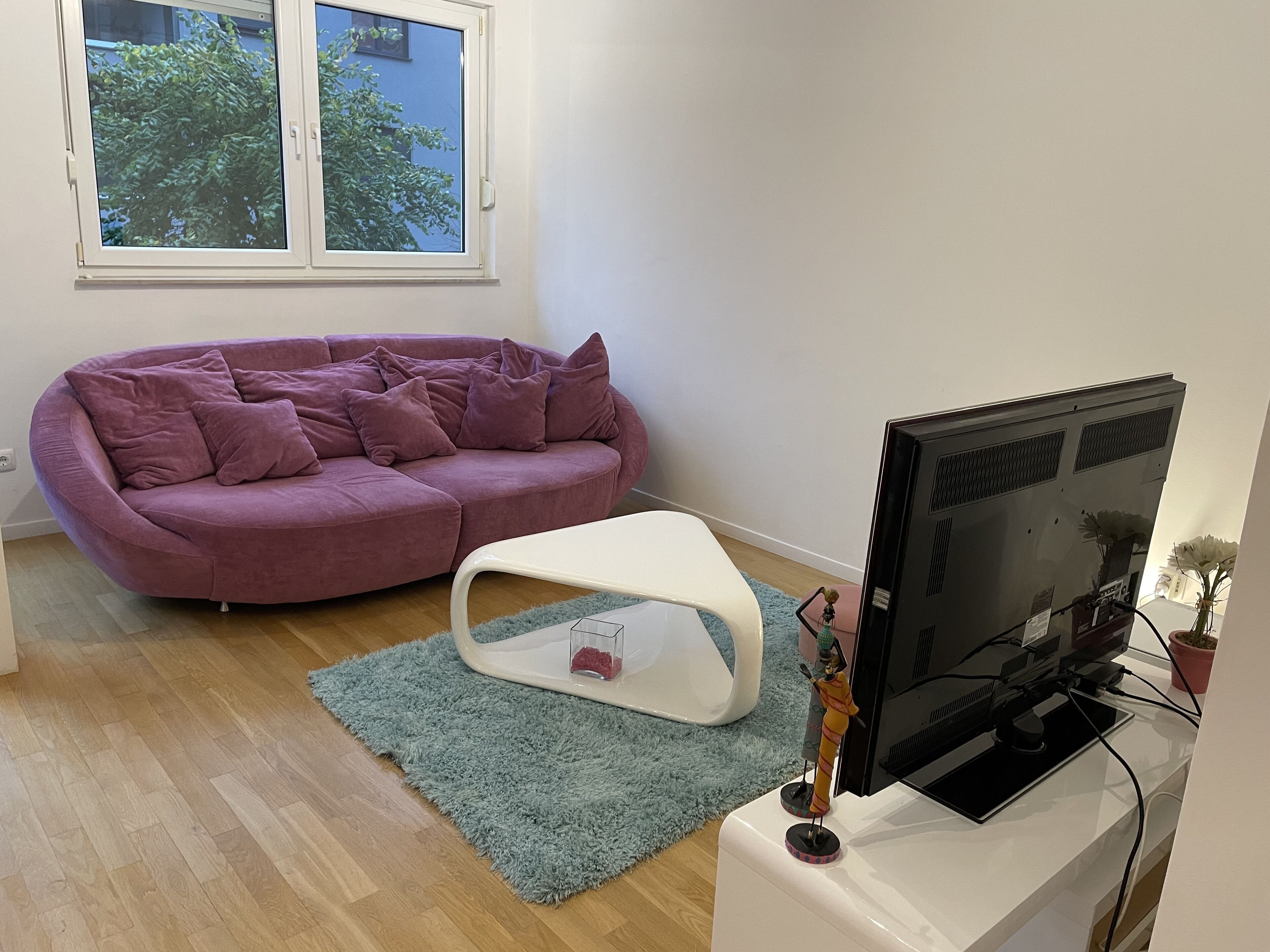 Un salon avec un canapé violet, un pouf blanc, un téléviseur à écran plat posé sur un meuble blanc et une fenêtre donnant sur des arbres.