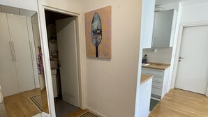 Una habitación amplia con piso de madera, un espejo grande, un cuadro en la pared y una cocina visible a través de una puerta abierta.