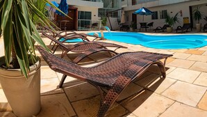 Zona junto a la piscina con sillones de descanso, un cielo azul despejado y un edificio de varios pisos al fondo.