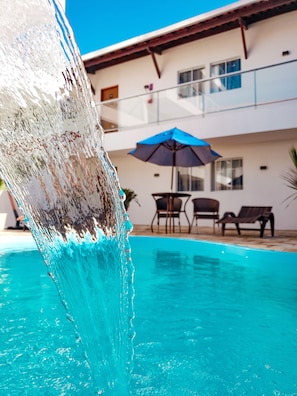 Una piscina con fuente de agua cristalina, una sombrilla azul y mobiliario exterior.