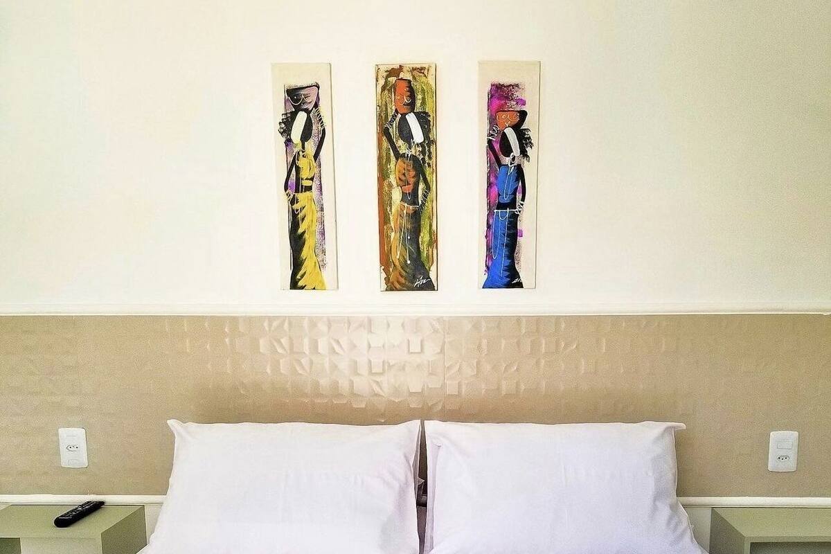 Een hotelkamer met een bed, witte kussens, een beige hoofdbord en drie kunstwerken aan de muur.