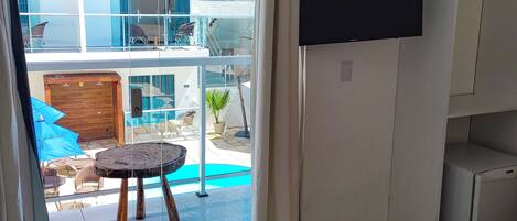A room with a large sliding glass door leading to an outdoor area with a pool, a flat-screen TV mounted on the wall, and a beige rug with two white pet beds.