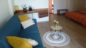 Un salon avec un canapé bleu, des coussins jaunes, une petite table basse ronde et un lit recouvert d’une couverture orange.