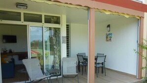 Un patio couvert avec une table et des chaises, une porte-fenêtre coulissante donnant sur un balcon, et un petit coin salon avec un canapé et un téléviseur.