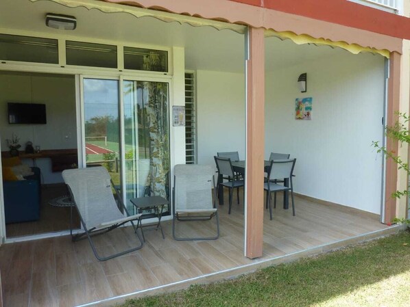 A covered patio area with a table and chairs, a sliding glass door leading to a balcony, and a small seating area with a sofa and a TV.