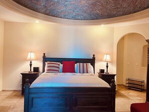 A bedroom with a large bed, two bedside tables with lamps, a brick-patterned ceiling, and a tiled floor.