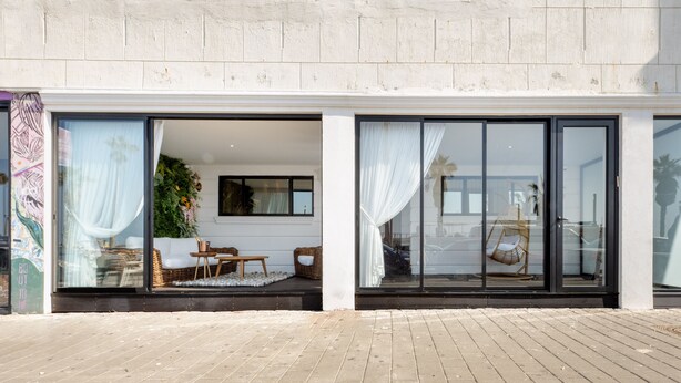 A modern building with large glass doors, a patio area with wicker furniture, and a view of the street. - Tripel-A (Tel Aviv)