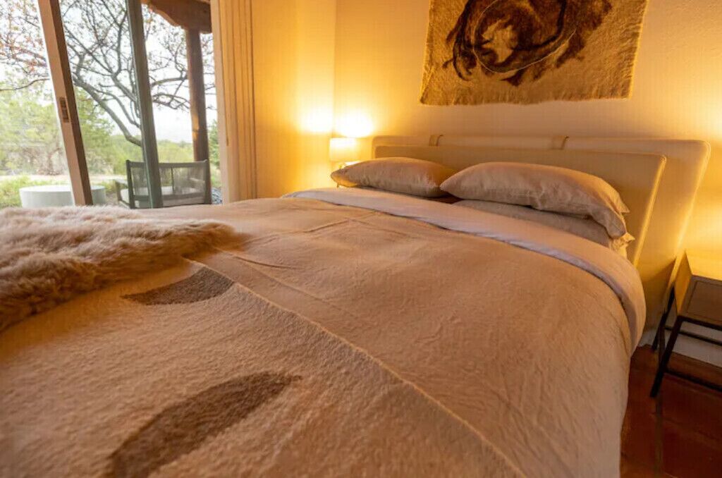 A bed with a white headboard, a fluffy white throw blanket, and two pillows. There is a large window with a view of trees and a chair outside.