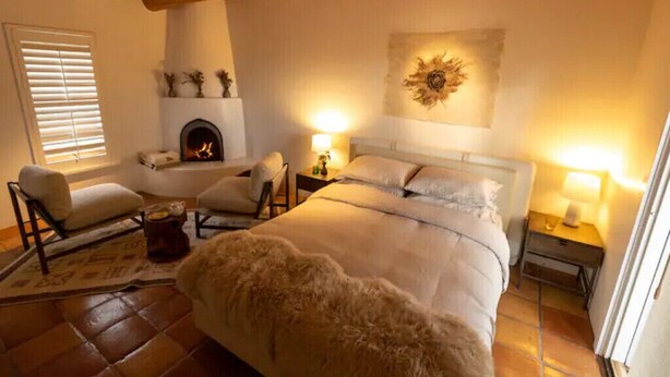 A cozy bedroom with a large bed, two chairs, a fireplace, and a nightstand with lamps. - Peaceful Suite in Desert Inn- Acoma Queen Room (Ojo Caliente)