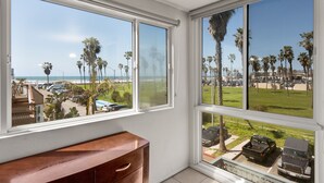 A room with large windows offering a view of a beach, palm trees, and parked cars. - Belmont Patio - Ocean View, Pet Friendly, Patio, & Garage (San Diego)