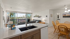 A modern kitchen with a large island, a dining area with wooden chairs, and a view of palm trees through large windows. - Belmont Patio - Ocean View, Pet Friendly, Patio, & Garage (San Diego)