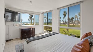 A bedroom with a large bed, a dresser, and a TV mounted on the wall. There are large windows offering a view of palm trees and a beach. - Belmont Patio - Ocean View, Pet Friendly, Patio, & Garage (San Diego)