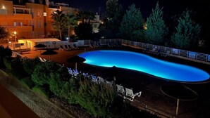 A nighttime view of a swimming pool with lounge chairs and umbrellas, surrounded by greenery and buildings.