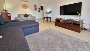 A modern living room with a grey sectional sofa, a light grey sofa, a wooden TV stand with a flat-screen TV, and a wall-mounted clock.