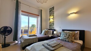 A hotel room with two beds, a ceiling fan, a balcony with chairs, and a wall-mounted light fixture.