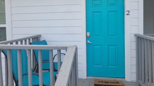 A white building with a turquoise door, a number '2' on the wall, and a wooden deck with blue cushions.
