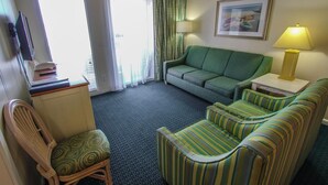 Ein Hotelzimmer mit einem grünen Sofa, einem gestreiften Sessel und einem Holzhocker. Es gibt einen Fernseher auf einem Schrank, eine Lampe auf einem Beistelltisch und ein gerahmtes Bild an der Wand.