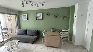 A living room with a grey sofa, a wooden dining table with chairs, a small round table with a wireframe base, and a green wall with framed pictures and hexagonal wall decorations. - Charming apartment in Sète close to all amenities, secure parking. (Sète)