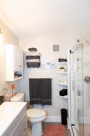 A bathroom with a toilet, sink, and shower. There are towels and toiletries on shelves. - Private Ground Level 2BR Condo (Frederick)