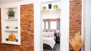 A room with a brick wall, a bed with white bedding, a bedside table, and a window with curtains. - Private Ground Level 2BR Condo (Frederick)