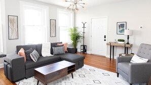 A modern living room with a sectional sofa, a coffee table, and a side chair. There is a coat rack, a potted plant, and a framed picture on the wall. - Private Ground Level 2BR Condo (Frederick)