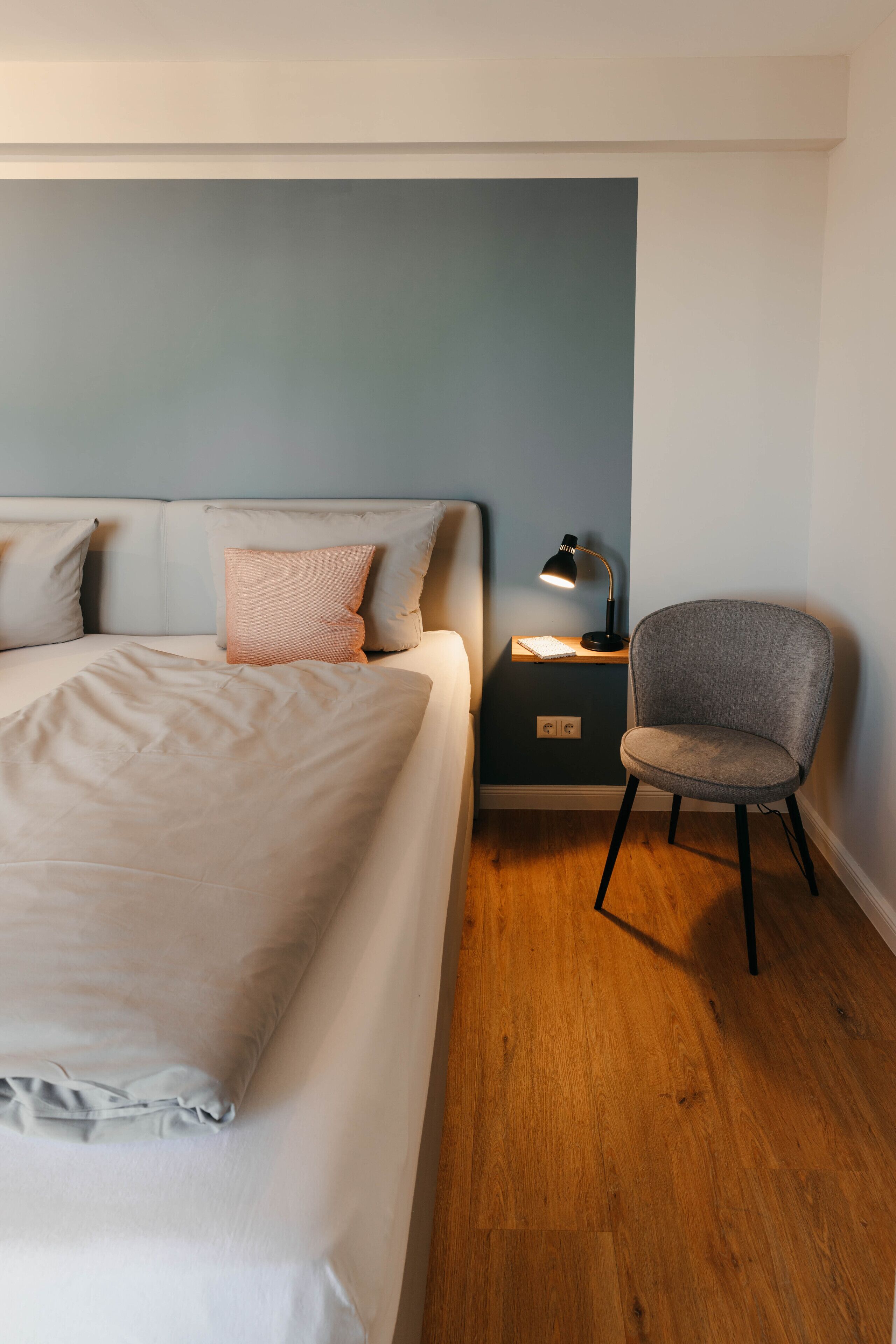 A bedroom with a bed, pillows, a chair, a nightstand, and a desk lamp.