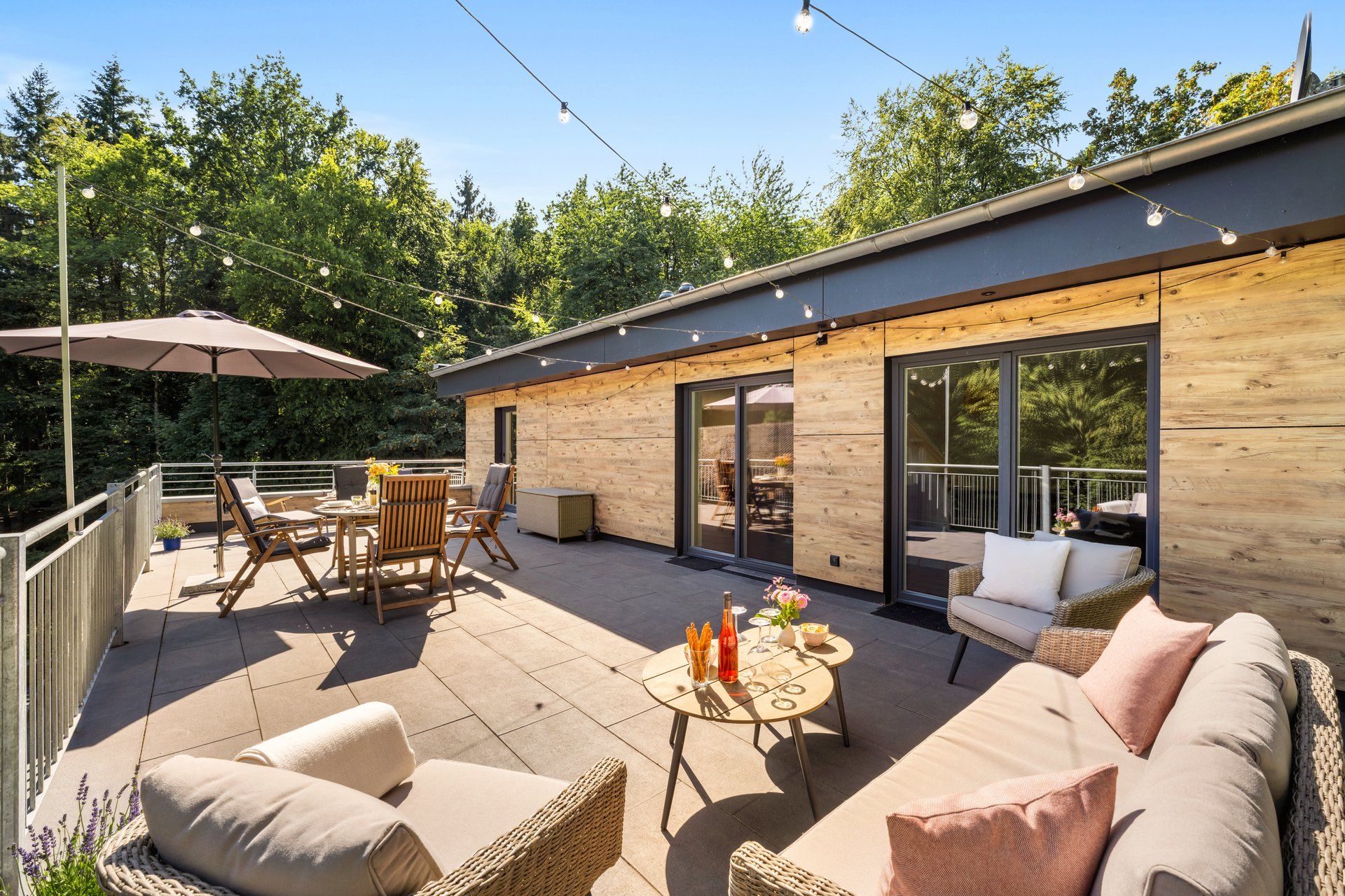 Un espace extérieur aménagé en terrasse, avec des sièges, une table où sont servis des boissons, et un bâtiment en bois doté de portes vitrées.