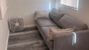A corner room with a grey sofa, a standing fan, and a window with blinds. - Cozy LBC house (Long Beach)