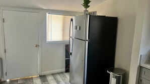 A compact kitchen with a black refrigerator, a trash bin, and a window with blinds. - Cozy LBC house (Long Beach)