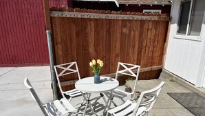 A patio with a round table and four white chairs, a wooden fence, and a small potted plant with yellow flowers. - Cozy LBC house (Long Beach)