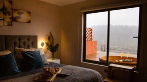 A bedroom with a large window, a bed with multiple pillows, a bedside table with a vase and a tray of food, and a view of a distant landscape. - Santo Domingo Lodge (Santo Domingo)