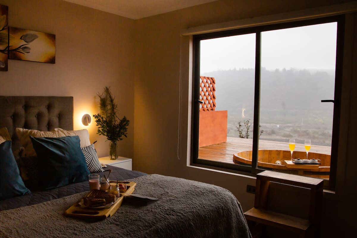 A bedroom with a large window, a bed with multiple pillows, a bedside table with a vase and a tray of food, and a view of a distant landscape.