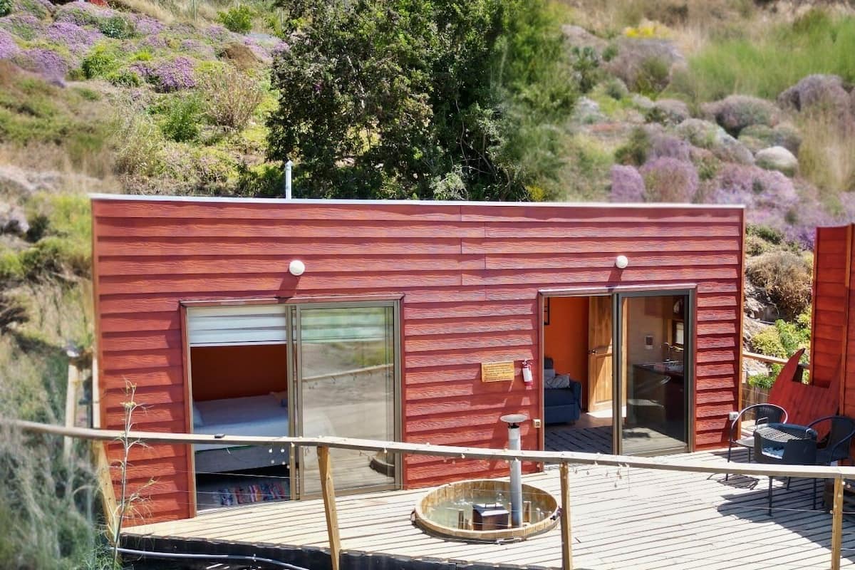 A modern cabin with a deck, large windows, and a view of the surrounding landscape.