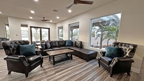 Ein geräumiges Wohnzimmer mit einem Ecksofa, Ledersesseln und einem Couchtisch. Deckenventilatoren und große Fenster sorgen für natürliches Licht.