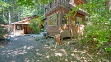 A cabin with a porch and a small deck, surrounded by trees and a gravel path.
