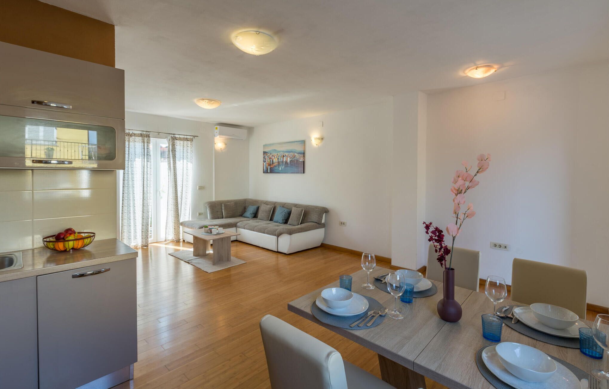 A modern living room with a gray sofa, a dining table set for two, and a kitchen area with a refrigerator and a fruit bowl.