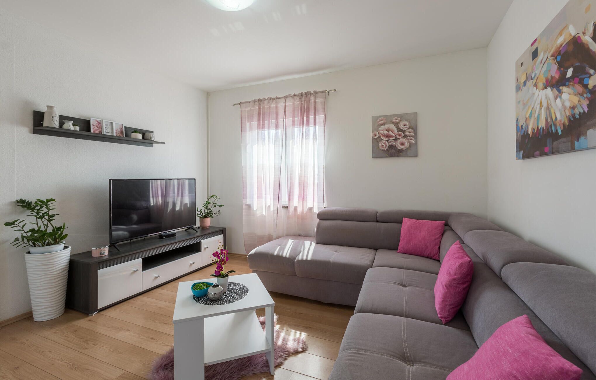 A modern living room with a grey sofa, a flat-screen TV, a white coffee table, and decorative plants.