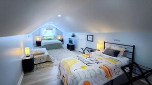 Ein gemütliches Schlafzimmer im Dachgeschoss mit schrägen Wänden, einem großen Bett mit Reise-Motivdecke, einem Schreibtisch mit Stuhl und einem Fenster mit Blick.